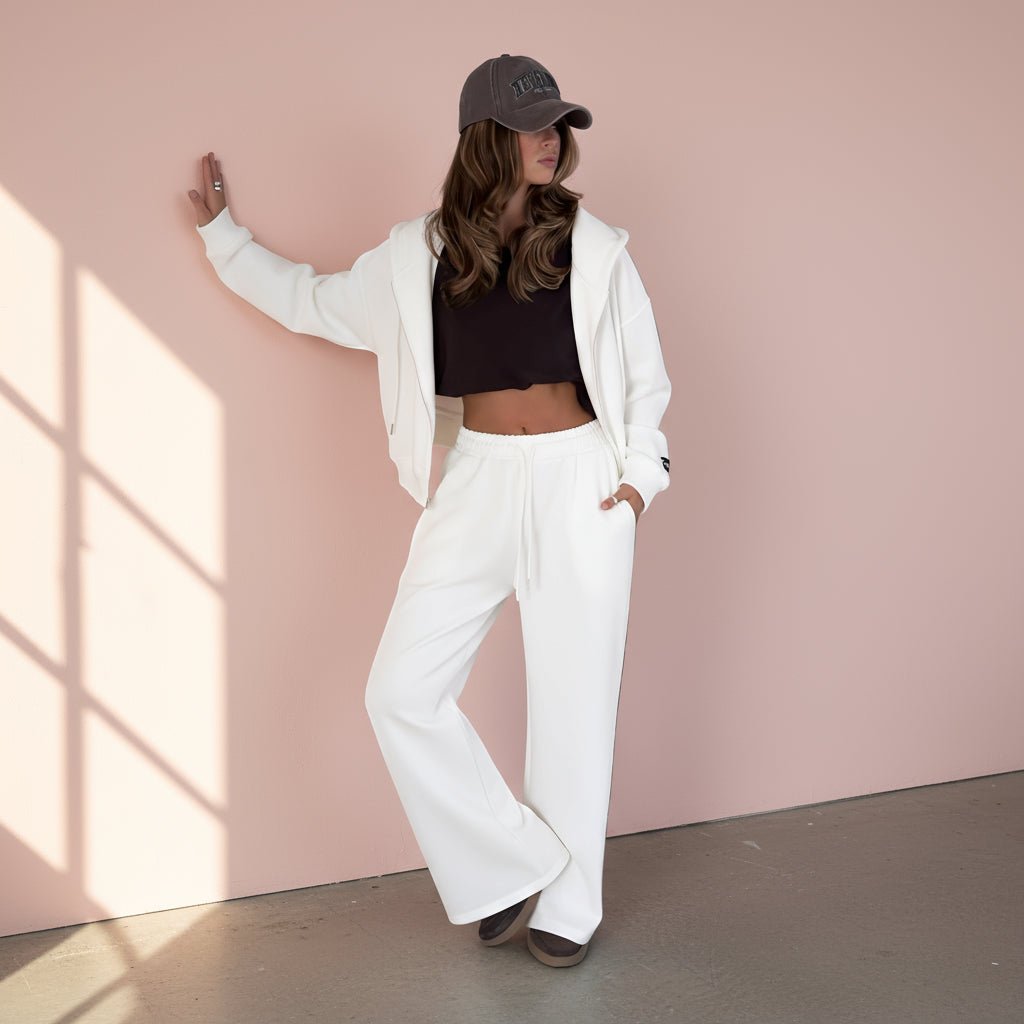 Person wearing a black crop top, white pants, and a white jacket with a gray cap on a light gray background
