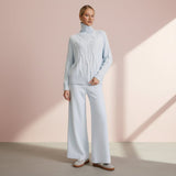 Woman wearing a light blue cable knit sweater and white pants against a pink background