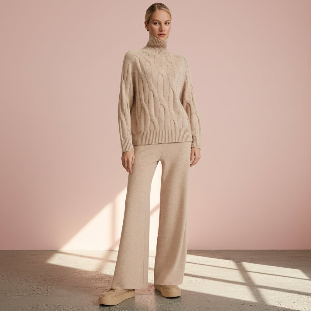Woman wearing a beige cable knit sweater and pants against a pink wall.