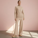 Woman wearing a beige cable knit sweater and pants against a pink wall.