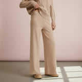 Person wearing a beige sweater and wide-leg pants against a pink wall.