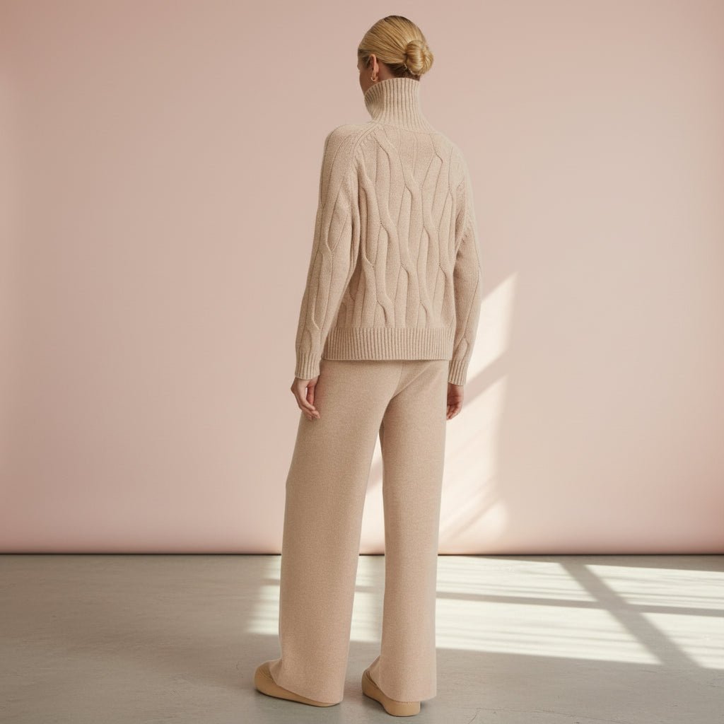 Person wearing a beige cable knit sweater and pants against a pink wall.