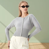 Woman wearing a light gray cardigan and white pants against a green wall.