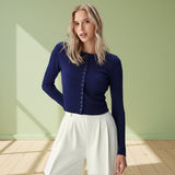 Woman wearing a blue cardigan and white pants against a light green wall.