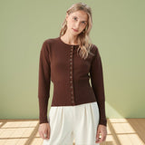 Woman wearing a brown cardigan and white pants against a light green wall.