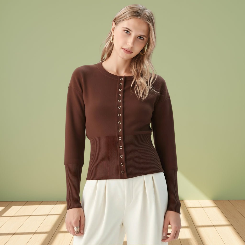 Woman wearing a brown cardigan and white pants against a light green wall.