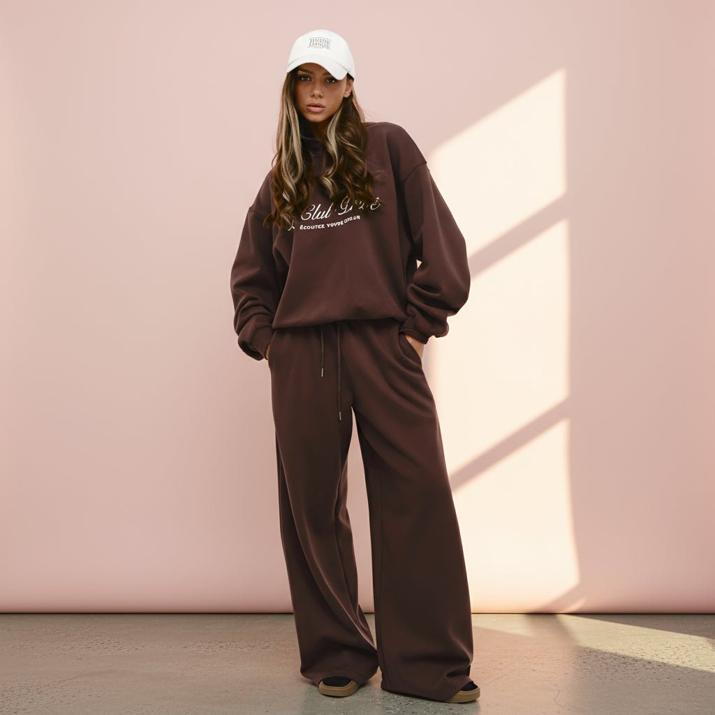 Person wearing a brown tracksuit and white cap against a pink wall