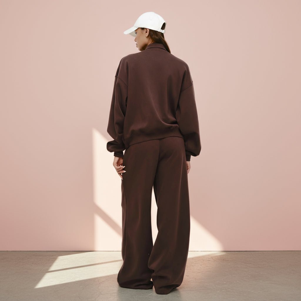 Person wearing a brown tracksuit and white cap against a pink wall