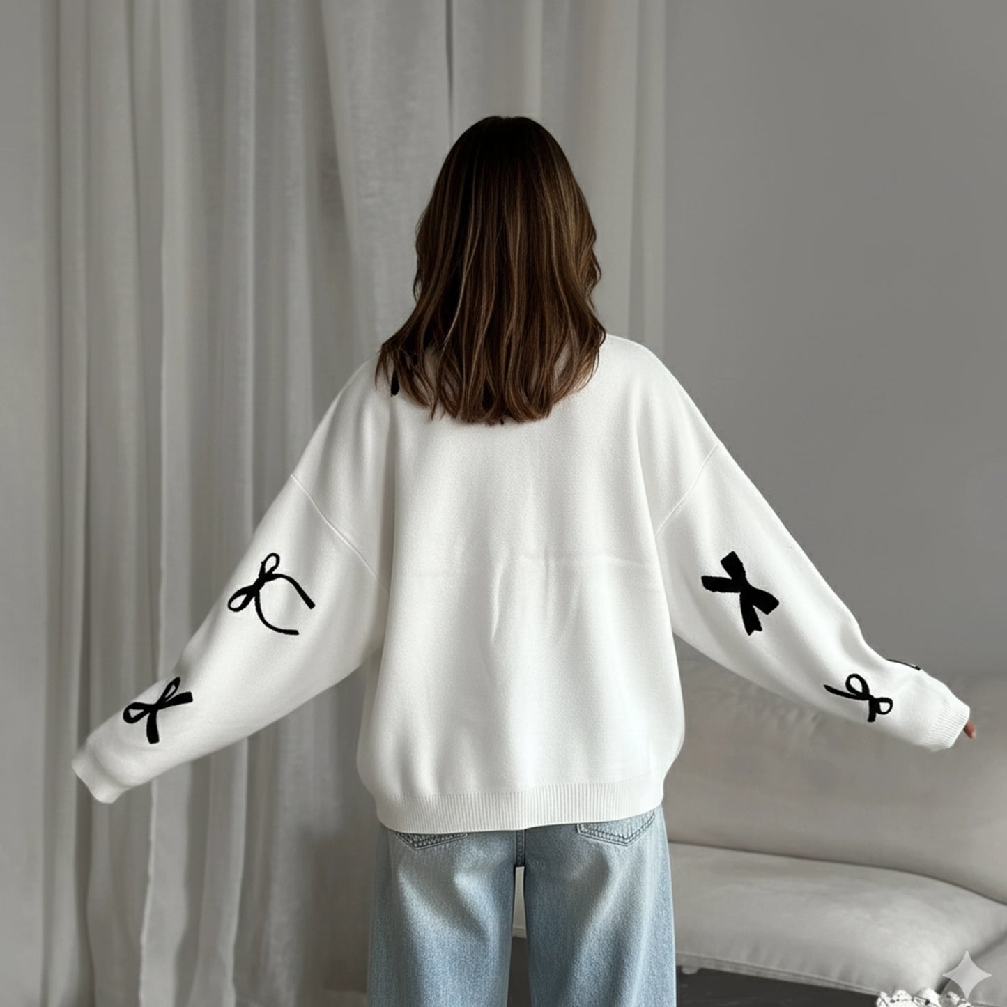 Person wearing a white sweater with black bow designs in front of a gray curtain.