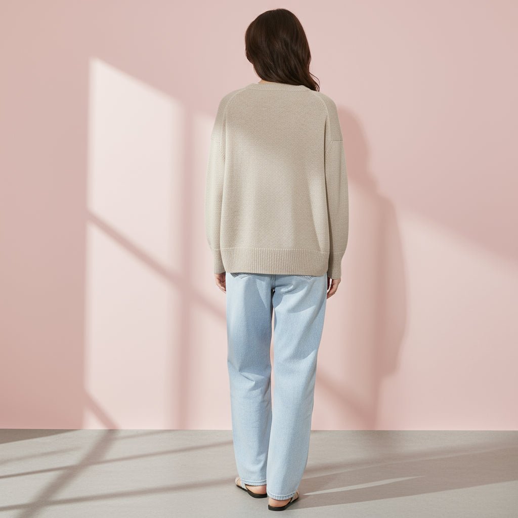 Person wearing a beige sweater and light blue jeans against a pink wall.