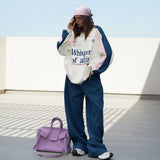 Person wearing a white sweatshirt with text and blue pants, holding a purple handbag on a light gray background