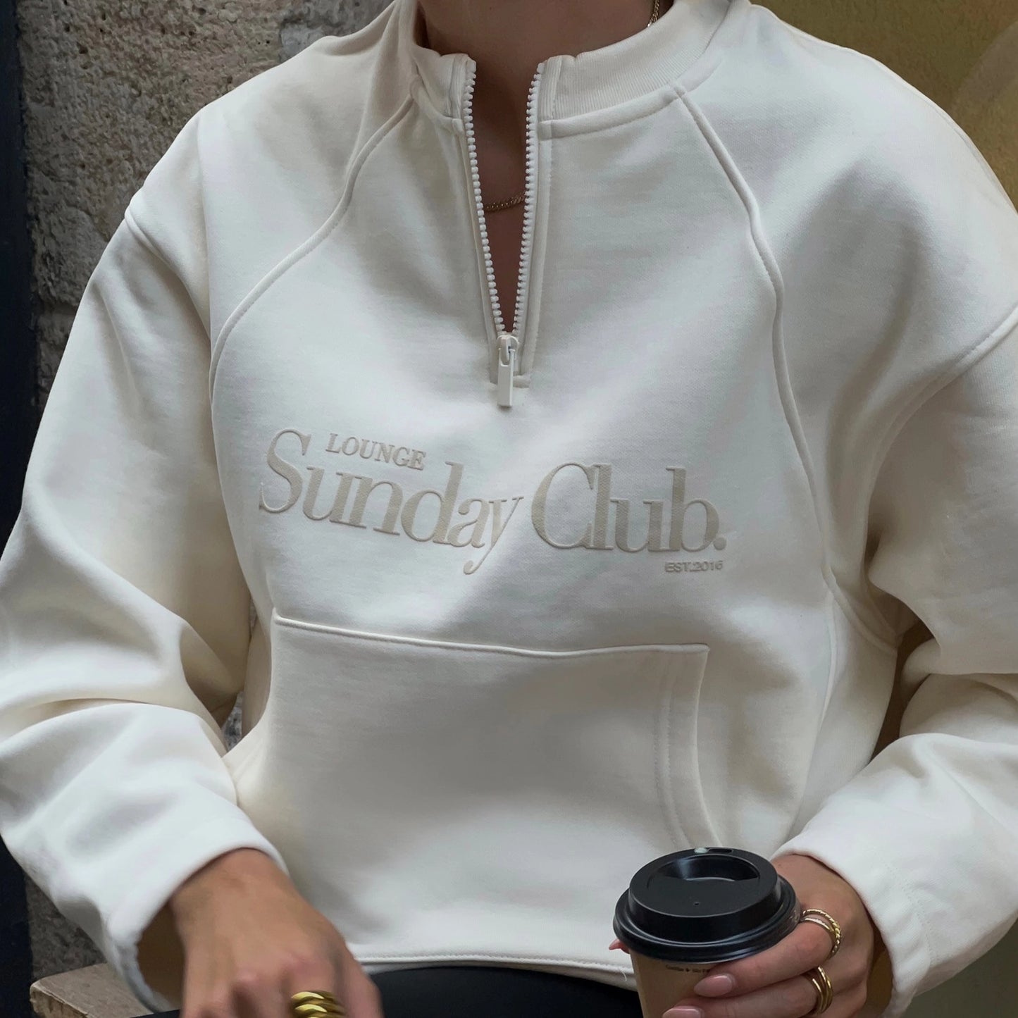 Person wearing a white hoodie with 'Lounge Sunday Club' text, holding a coffee cup.
