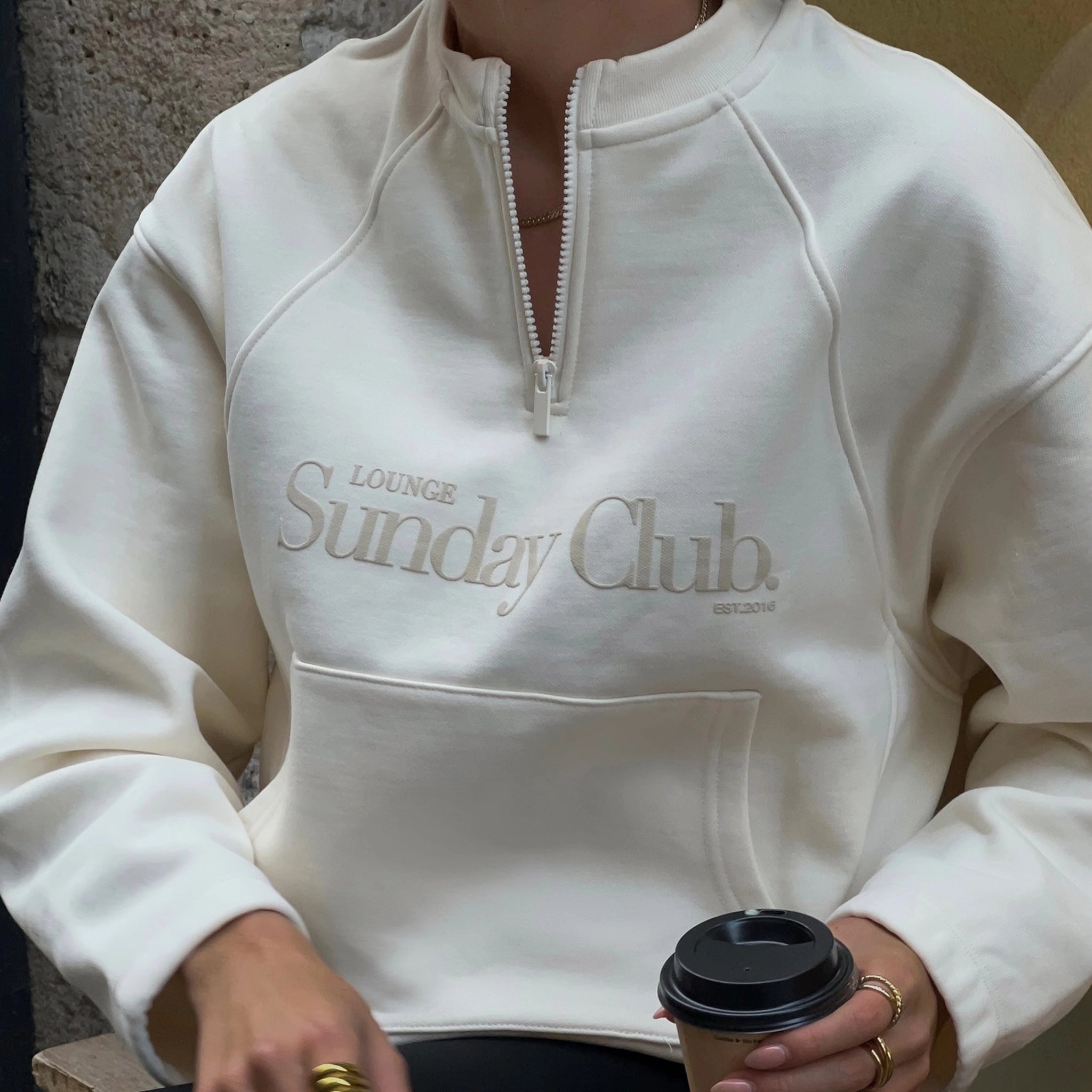 Person wearing a white hoodie with 'Lounge Sunday Club' text, holding a coffee cup.