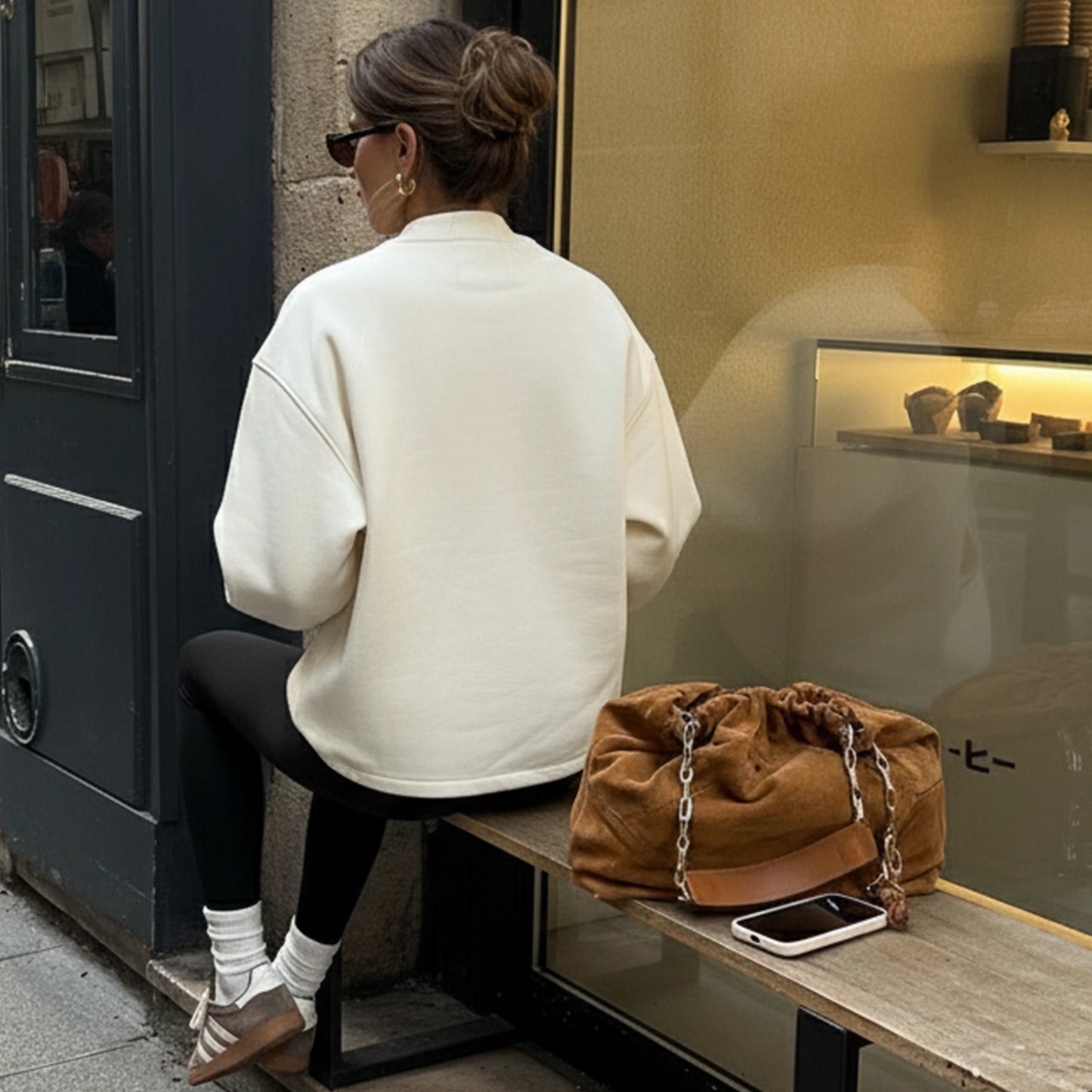 Woman sitting on a bench with a brown bag and phone next to her, in front of a store window.